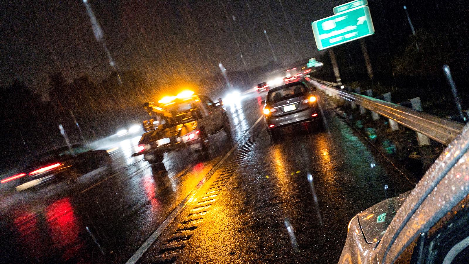 A tow truck's amber light bar and the hazard lights of a stopped car reflected on a rain-wet highway at night
