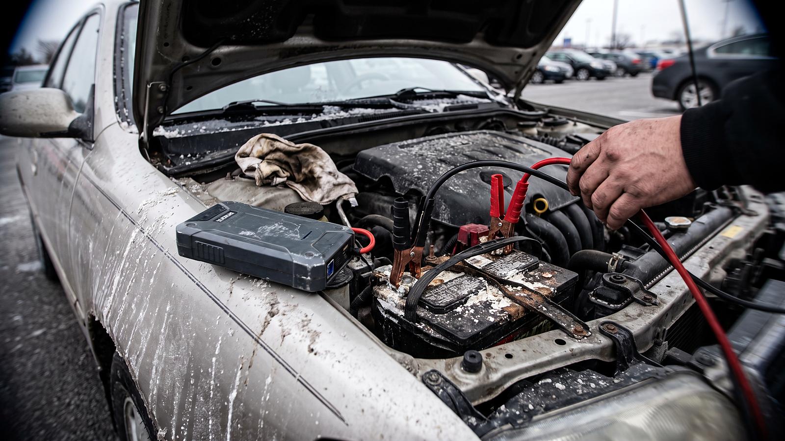 Jumper cable clamps on a car battery terminal under an open hood, with a portable jump pack resting on the fender